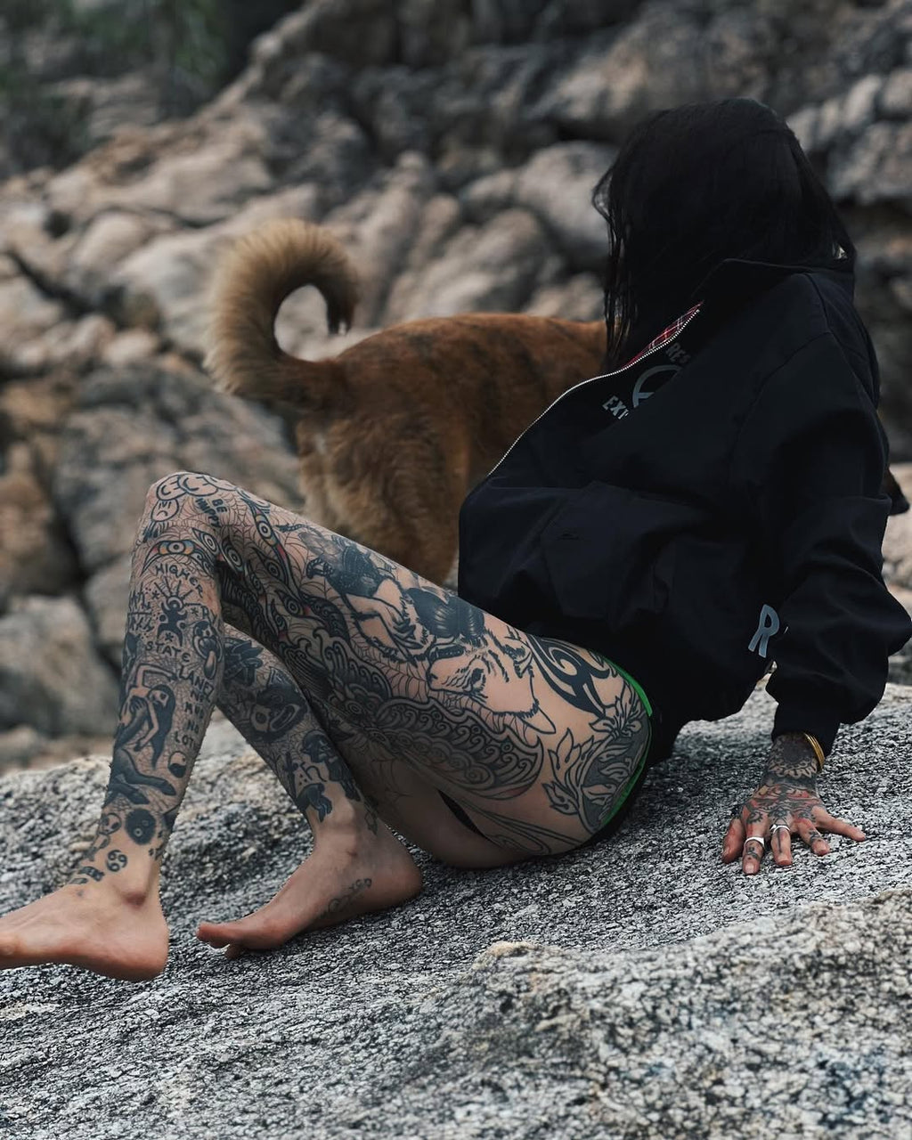 Person with tattoos sitting on rocks with a dog